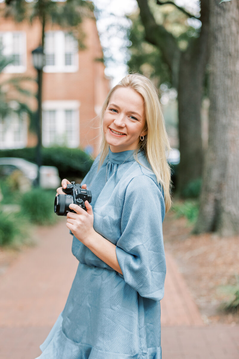 Newport RI Wedding Photographer | Deirdre wearing a light blue dress. She's standing on a brick path, with trees and brick buildings in the background.