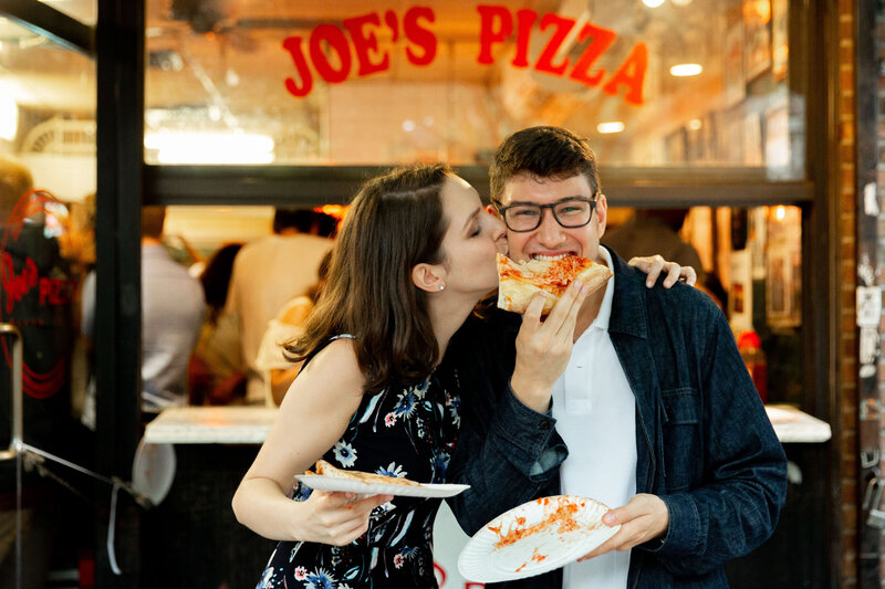 A person eating a piece of pizza while their partner kisses them on the cheek.