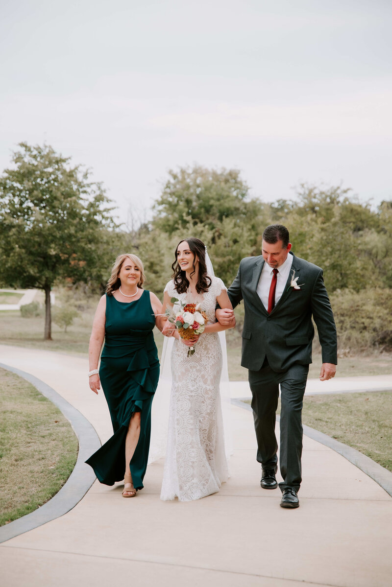 A bride, her mother, and the father of the bride are walking down a paved path.