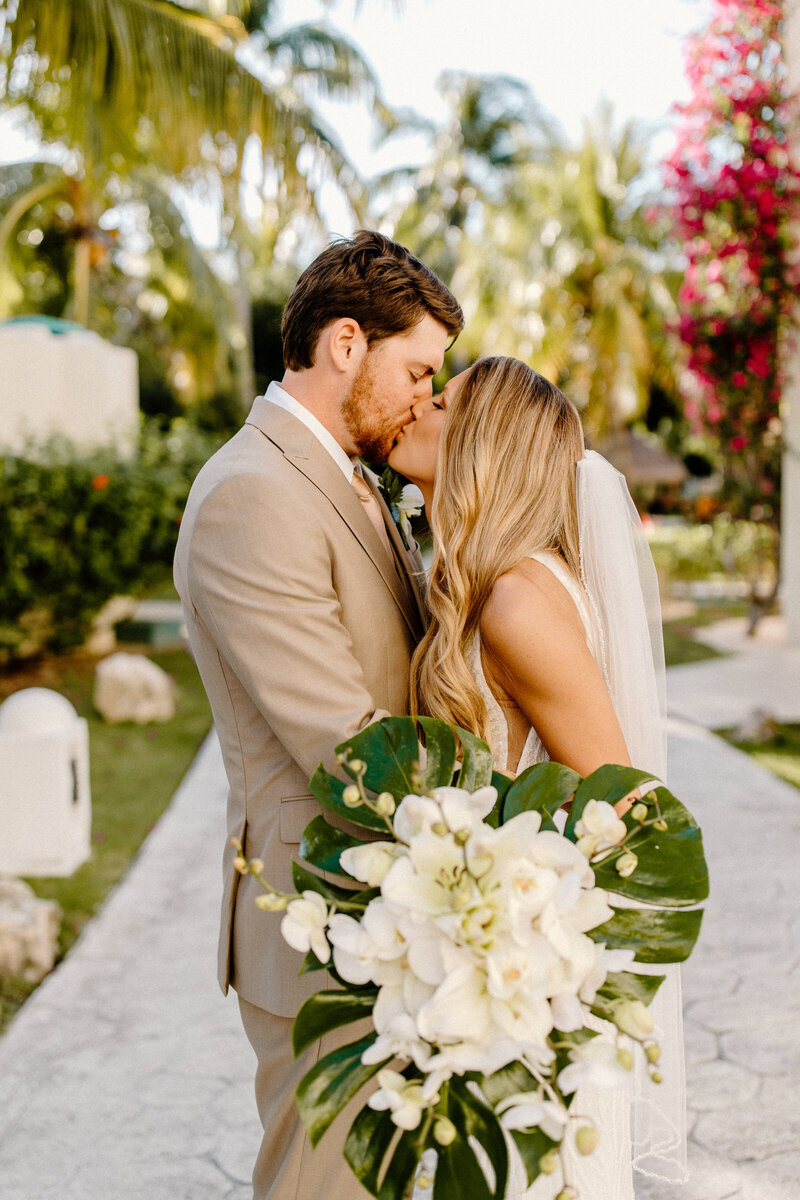 Smiling bride and groom leaning in for a kiss, holding a tropical bouquet with palm trees in the background in Cancun.