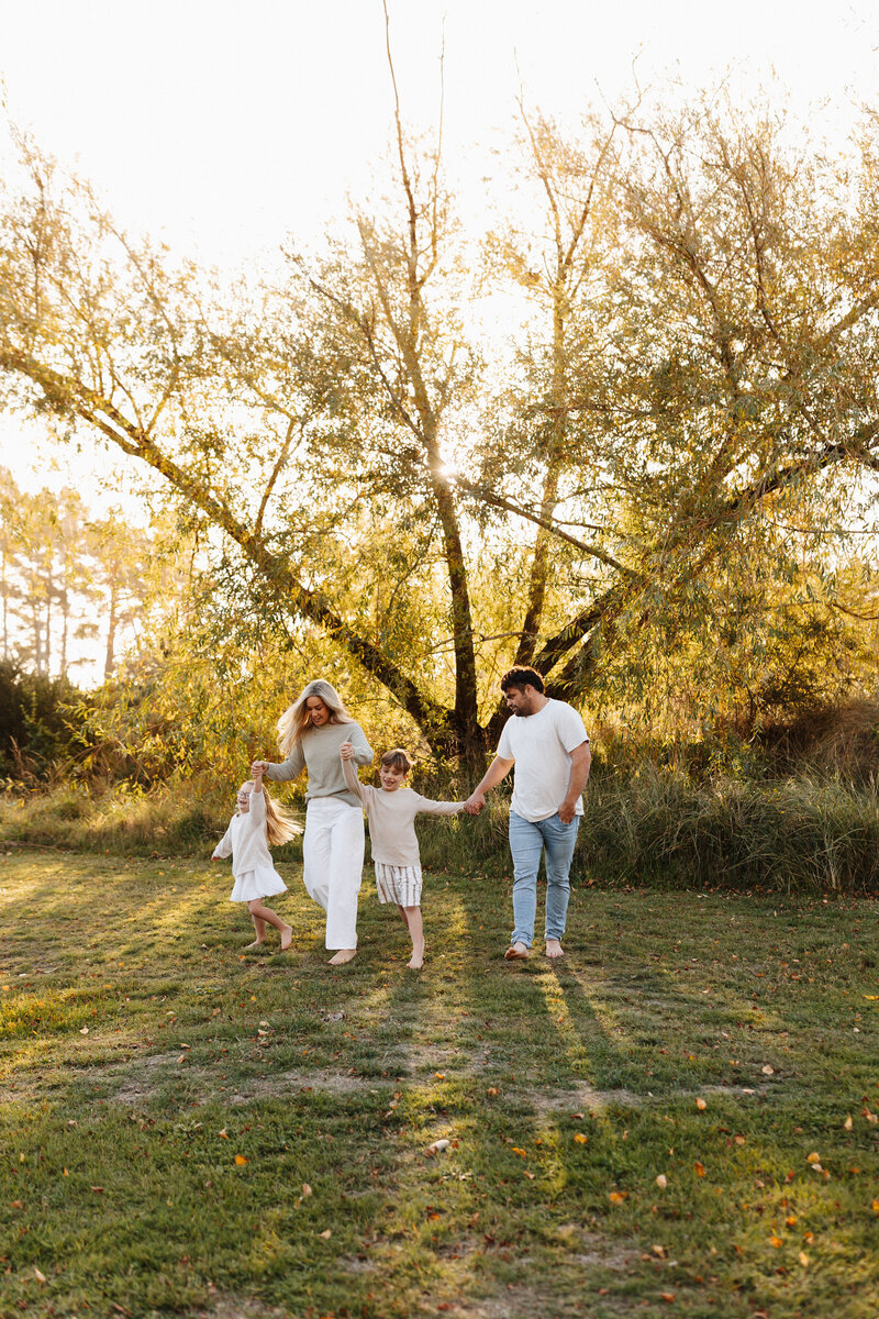 A mother and father hold their daughters hands and walk through a park while the sun sets behind them during a family photoshoot with canterbury photographer lucy jane