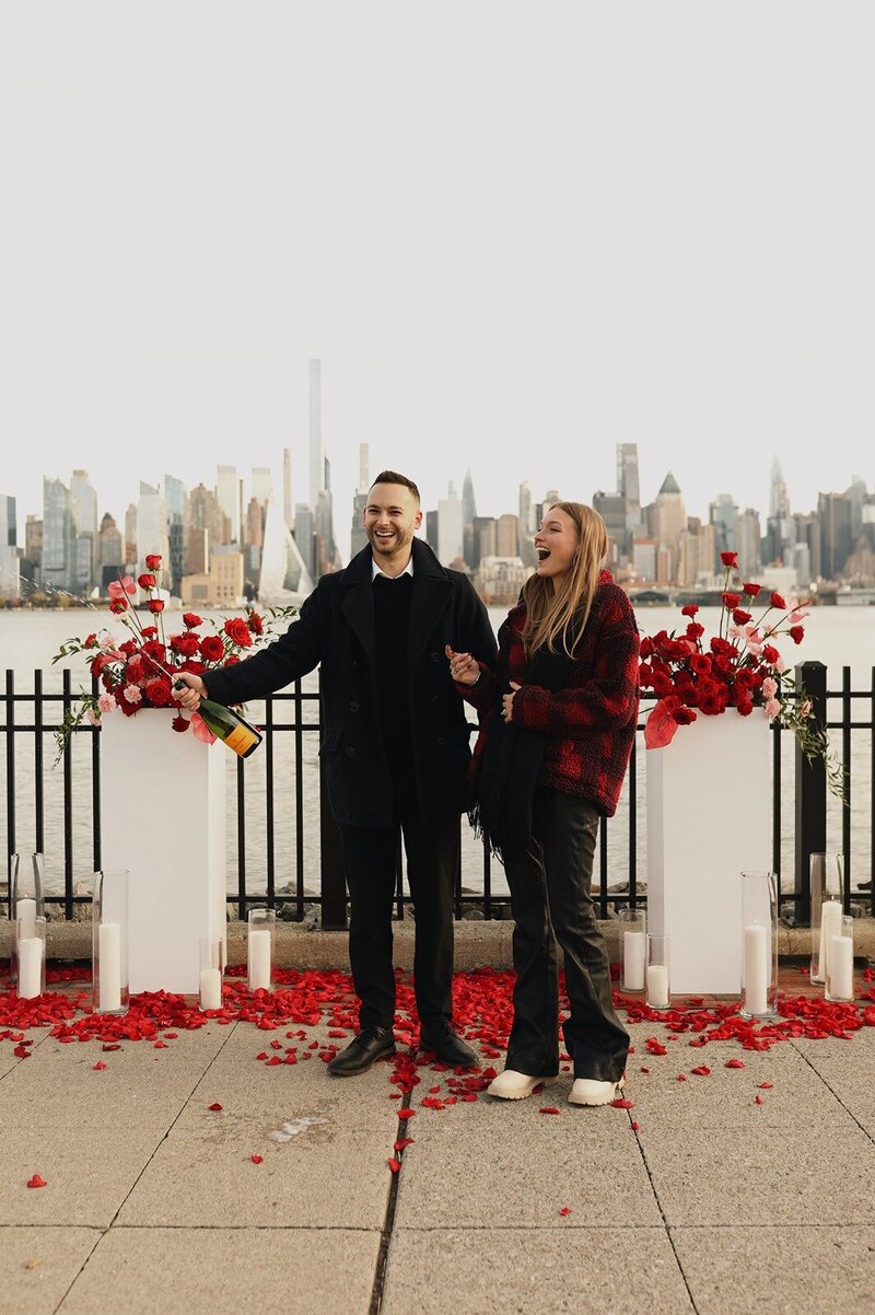Couple celebrating their engagement on a rose-petal-strewn waterfront with stunning New York City skyline views, champagne in hand and red floral arrangements framing the scene. Styled by Yours Truly, a wedding planner and designer in New Jersey.