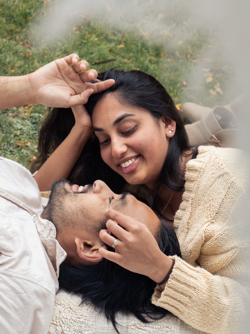 a couple celebrates their engagement with an autumn picnic in Hagley Park in Christchurch, New Zealand. they are laying on a blanket and gently caressing each other's faces and smiling. the woman has an engagement ring visible on her left hand.