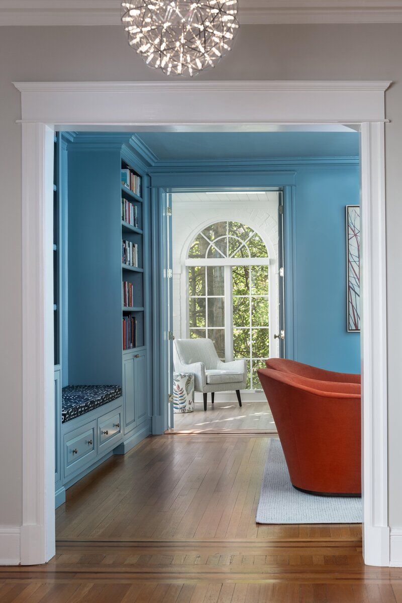 Home library featuring blue walls, built-in bookshelves, an arched doorway leading to a sunlit room, and an orange chair