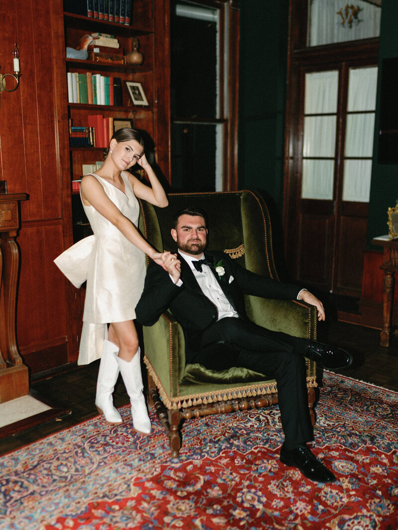 Bride in short dress and boots leaning against groom sitting in green velvet chair