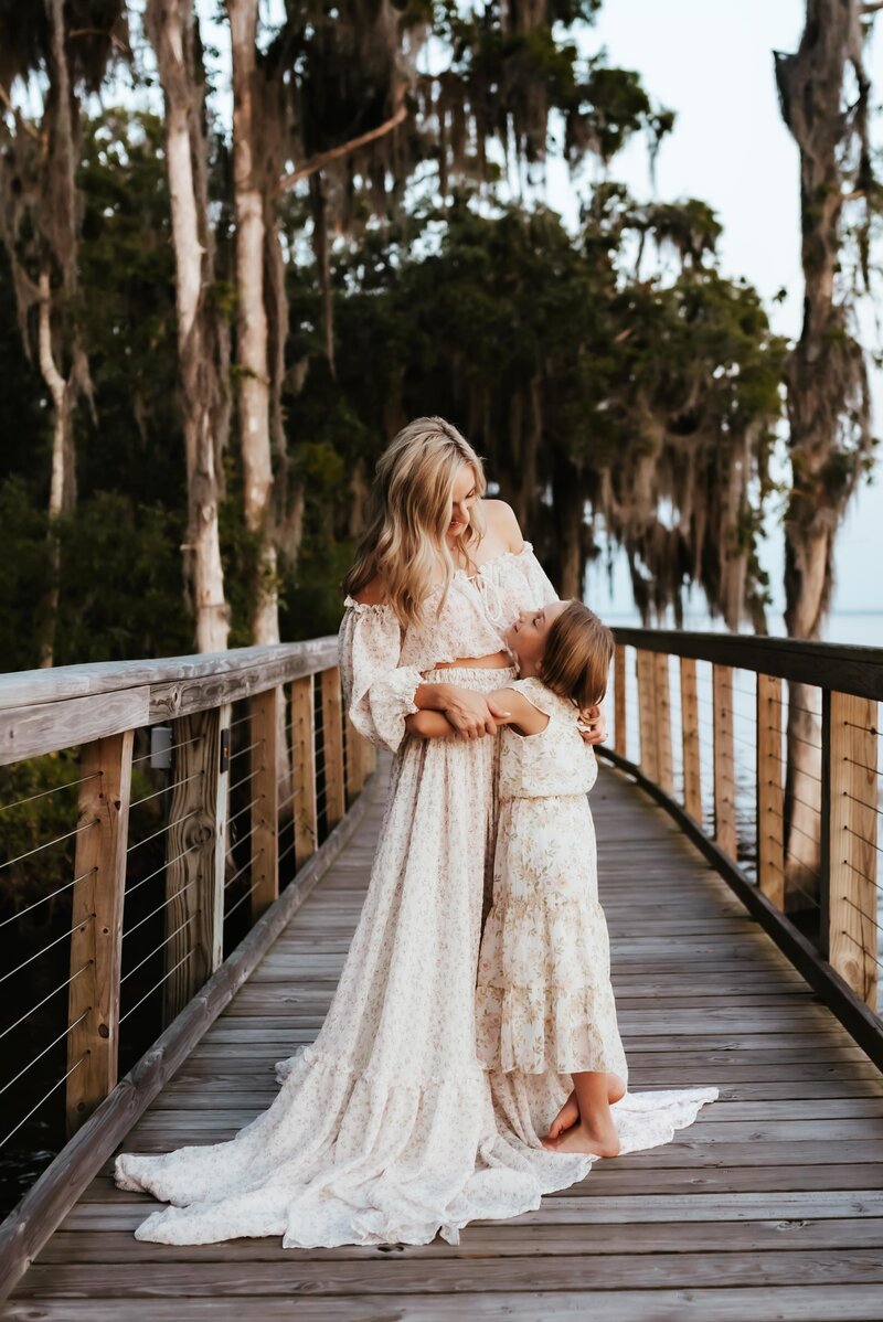 A mother in a long white dress embracing her daughter on a board walk.