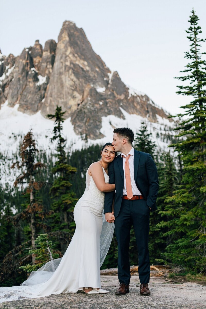The golden orange alpenglow shines over Broken Top as an Asian Bride wearing a white v-neck gown holds her groom hand walking along the creek at their Sparks Lake Oregon Elopement near Bend Oregon. | Erica Swantek Photography