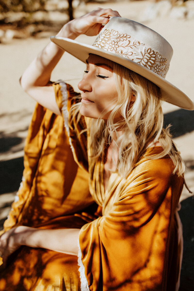 Family and Maternity Photographer,  Kylie sits reflectively as she holds her hat to her head in the sunlight, her blond hair falls onto her shoulders and an auburn  outfit