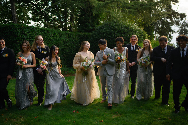 A joyful wedding party walks together on a lush green lawn, surrounded by tall hedges and trees. The bride, in a whimsical gold gown adorned with delicate details, and the groom, in a light gray suit, share a laugh at the center. Bridesmaids in shimmering silver dresses and groomsmen in classic black suits complement the scene, holding vibrant floral bouquets. The atmosphere is lighthearted and full of celebration, capturing the beauty of an outdoor summer wedding.