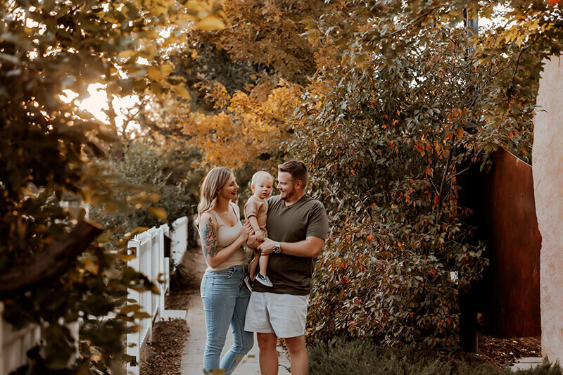 Family of three standing next to a white fence and fall trees on a warm day in downtown Lafayette Colorado
