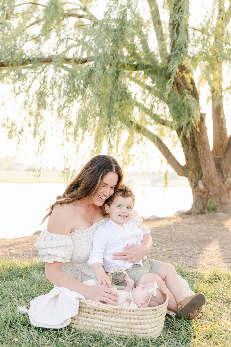 mother sits in front of willow tree with son on her lap looking down at her newborn laying in a moses basket, Central Indiana family photographer