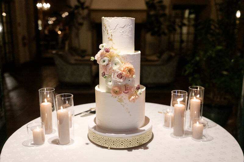 November wedding at Graylyn Estate, designed and planned by Charming Carolina — three-tiered white wedding cake with edible gold flecks, displayed on a white linen and surrounded by candles in the sun room overlooking the Atlantis Room.