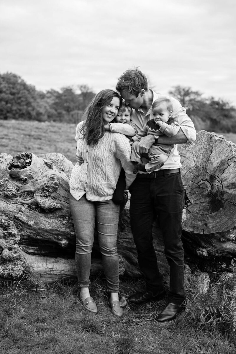 A mother gives her small son a piggy back while her husband kisses her holding his young son. Photographed by Jess Morgan during a photo shoot in Richmond Park