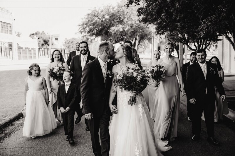 a bride and groom walk hand in hand while sharing a kiss. Their bridal party is walking behind them cheering them on while they walk the streets of downtown abilene, texas. 