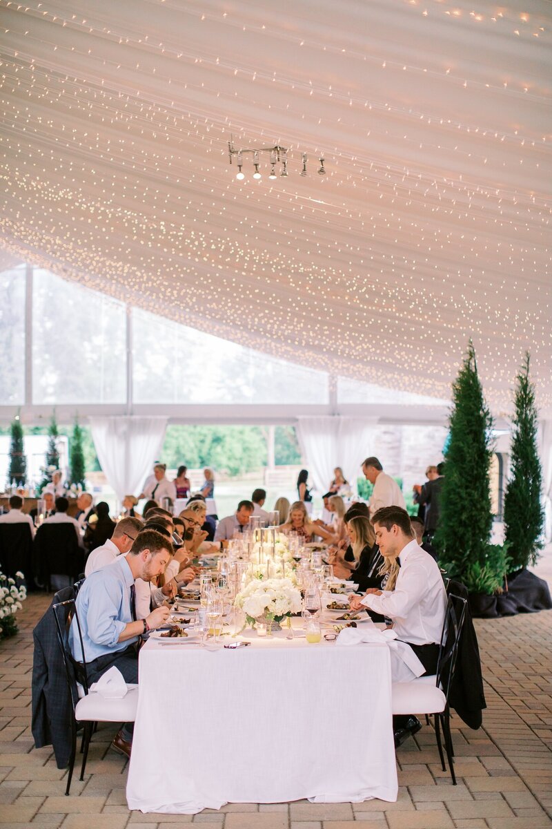 Guests dining under a tent with string lights and white table settings.