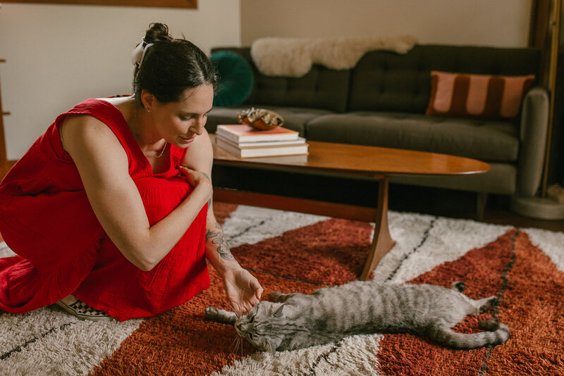 A woman in a red dress bending down to pet a gray cat who is laying on a carpet in front of a coffee table. 