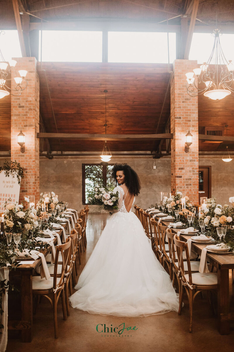 Bride walking through rustic reception setup with Crossback chairs, long banquet tables, white florals, and chandeliers overhead