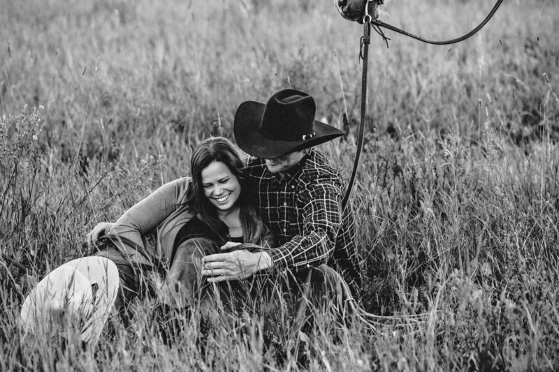 engaged couple sits in tall grass with cowboy hat and horse