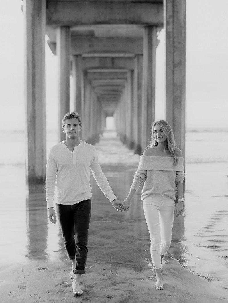 couple walking hand in hand smiling under the scripps pier in la jolla wearing jeans and knit white sweater