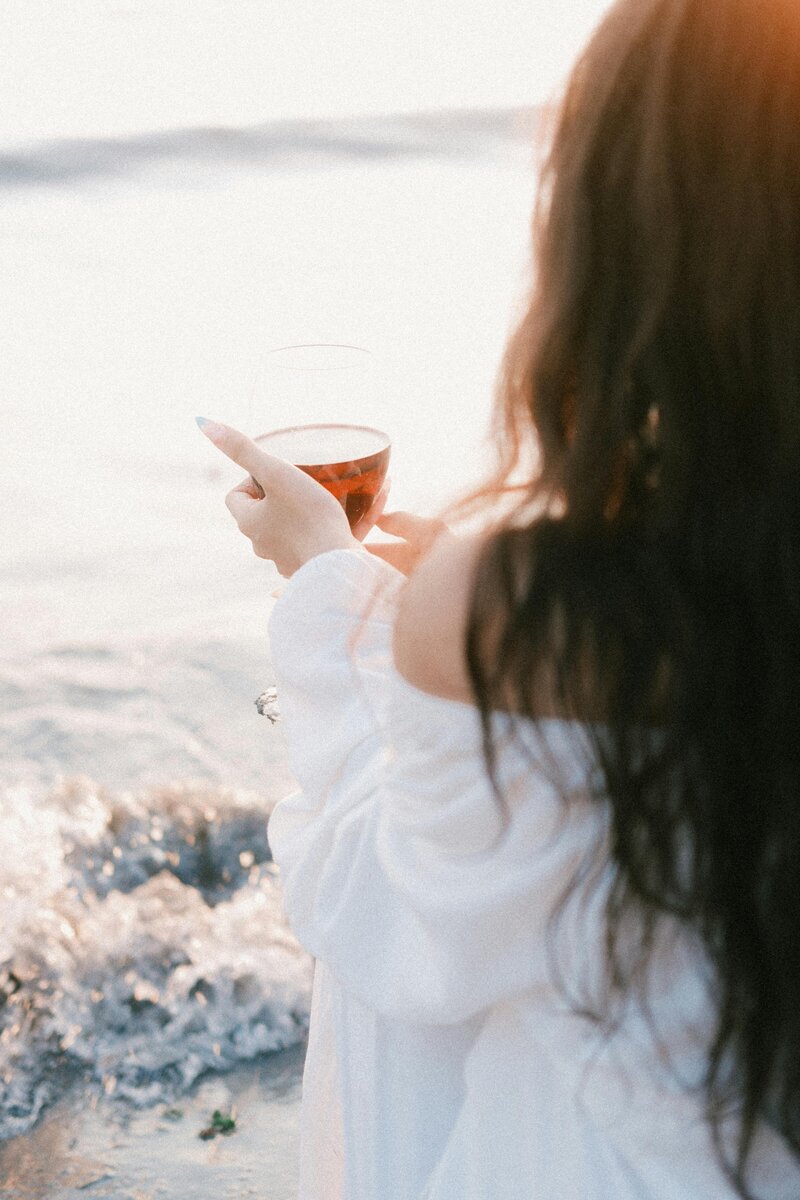 Woman with long wavy brown hair and a white dress holding a glass of wine looking into the ocean