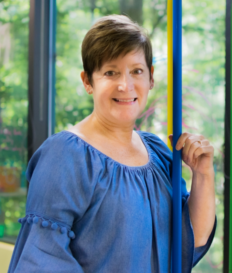 Michaelene is a middle aged woman with short pixie cut brown hair with hazel eyes and a kind smile. She is wearing a denim shirt with a scoop neck & flared sleeves.