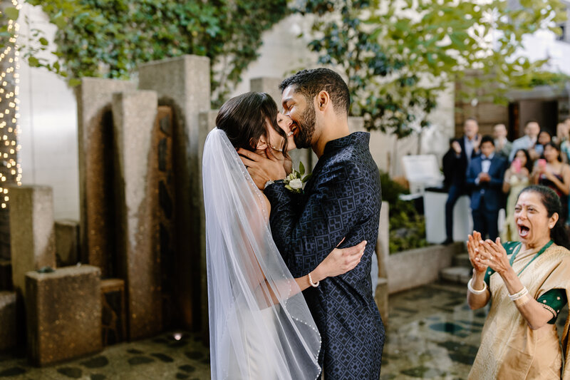 First kiss between bride and groom with mother of groom watching with enthusiasm