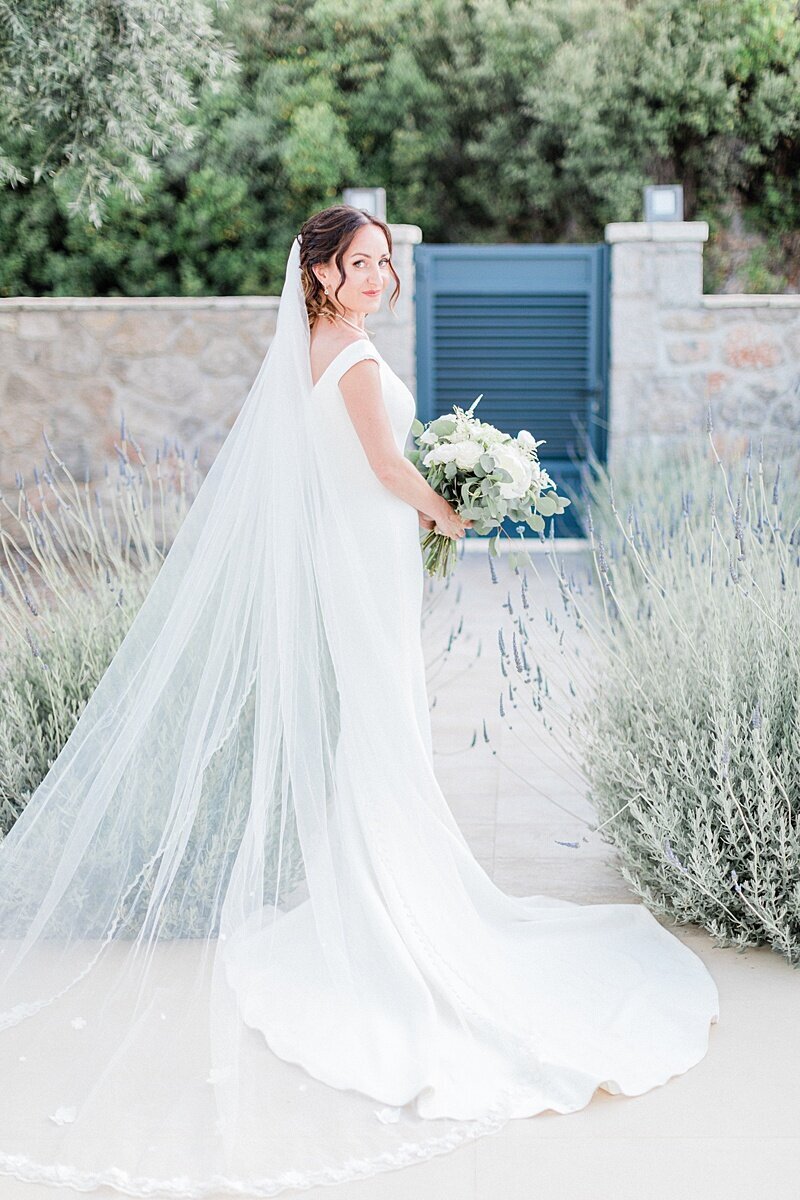Bride in a sleek white gown holding a bouquet, standing between lavender bushes with a soft veil flowing behind her in a serene garden setting

