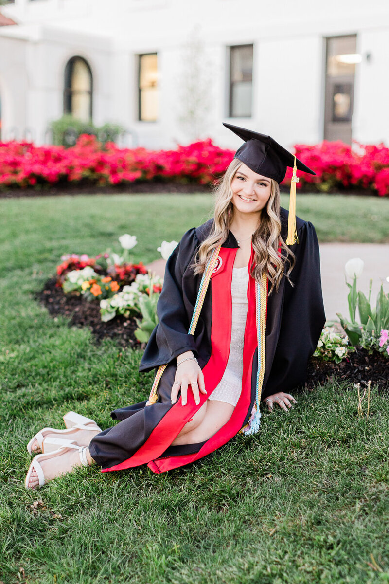 Girl sitting in the grass in a black cap and gown with red stoles