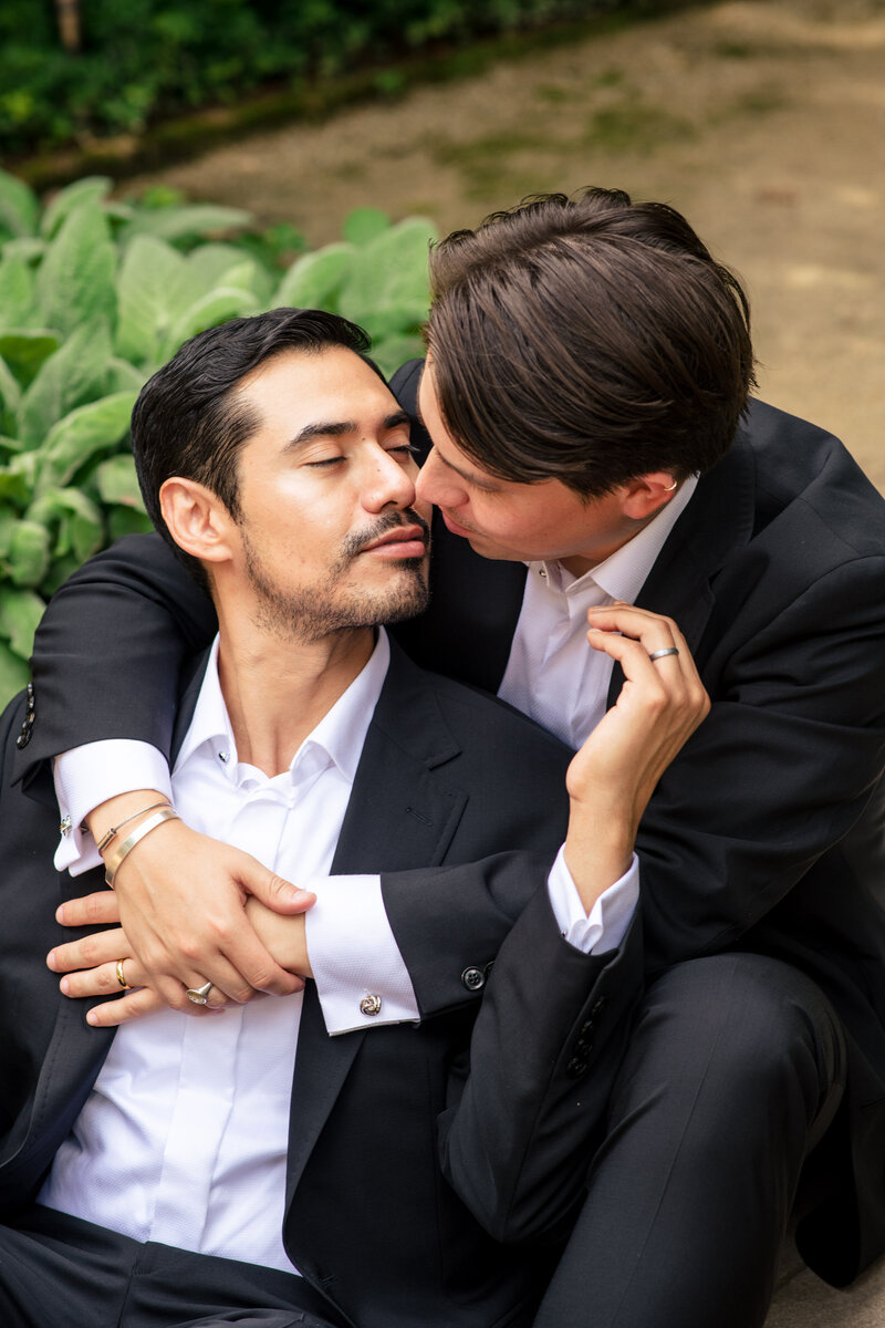 Photograph of a same-sex male couple seated and snuggled up about to kiss in the gardens at Duke Mansion  by Charlotte wedding photographers DeLong Photography