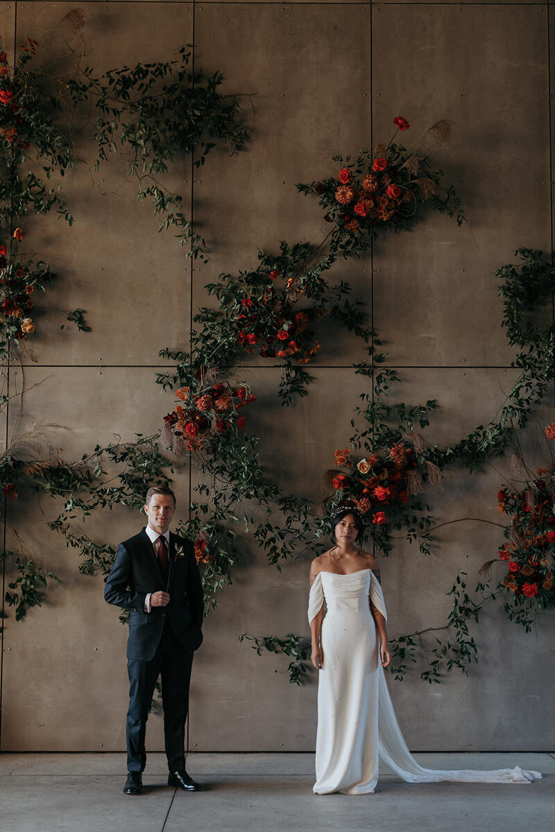 Catskills Groom and Bride pose in front of giant concrete wall with moody autumn botanical  design with deep pops of red and orange florals.