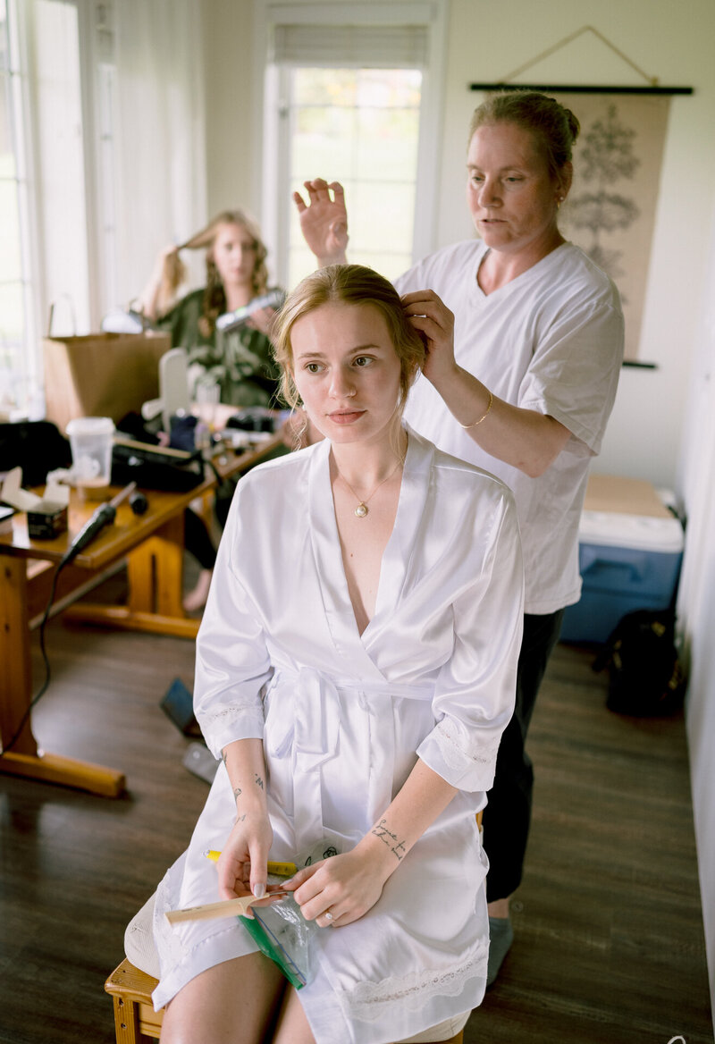 A woman getting her hair styled while holding a brush, with another woman working on her hair, in a room with makeup items on the table.