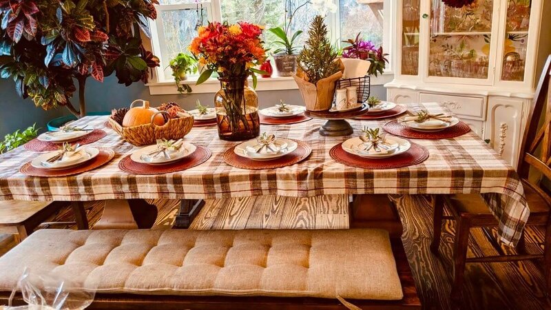 Festive dining table set with a plaid tablecloth, autumn-themed centerpiece, and matching plates with decorative napkins. Surrounded by plants, theres a cushioned bench and basket of fruit, under a sunny window.
