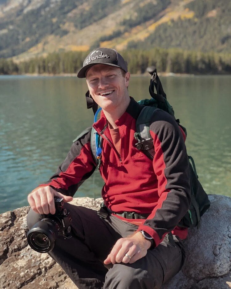 Elopement Photographer poses with camera and hiking bag at a lake