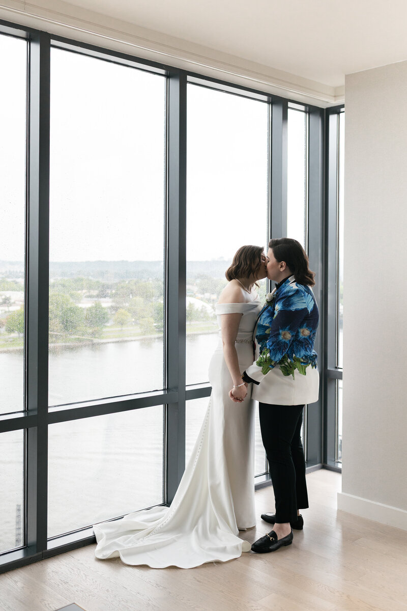 lesbian brides kissing in front of a floor to ceiling window at the Wharf in DC overlooking the water