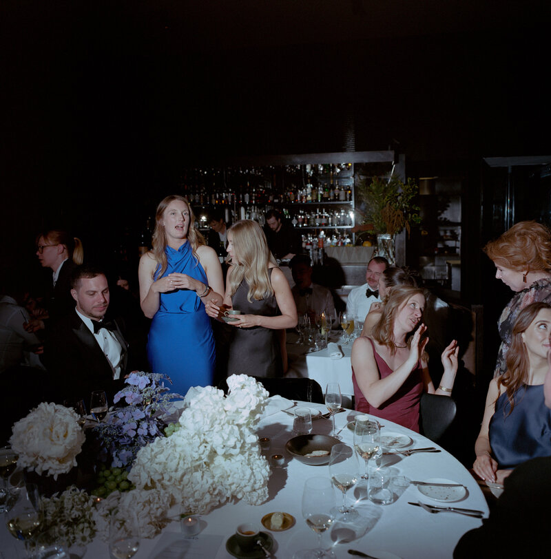 A vibrant wedding reception scene at Cutler and Co in Fitzroy, Melbourne, capturing a mix of lively conversations and relaxed moments among the well-dressed guests. The round table in the foreground is adorned with a lush centerpiece of white hydrangeas and purple flowers, with clusters of green grapes adding a natural touch to the elegant decor. The table is set with empty wine glasses, water glasses, and scattered plates, indicating that guests are mid-reception, enjoying themselves after dinner.  In the center, two women stand out, one in a striking blue dress and the other in a sleek dark gown, engaged in cheerful conversation. Surrounding them are seated guests in formal attire, with one woman in a burgundy dress smiling and gesturing animatedly to someone off-camera. The men, dressed in black-tie suits, contribute to the refined atmosphere. Behind the group, a well-stocked bar is visible, where bartenders are preparing drinks, adding to the lively yet intimate ambiance. The lighting is soft and warm, creating subtle shadows that accentuate the elegant details of the room, while the dark background allows the colorful clothing and decor to pop. The atmosphere at this wedding reception is one of joy, elegance, and celebration, perfectly suited to the upscale venue of Cutler and Co.