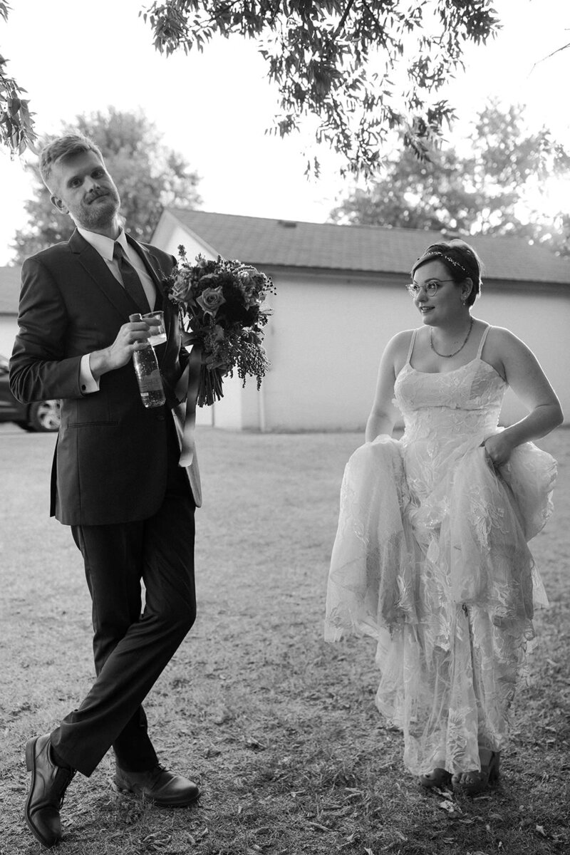 Black and white image of a couple during their wedding day, with the groom holding a drink and the bride lifting her dress slightly