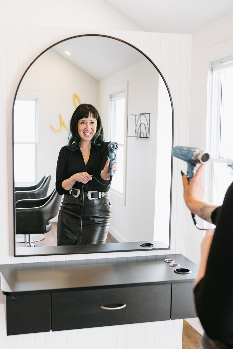 A mirror shot of a salon hair stylist pointing a hairdryer at her reflection for a team branding photoshoot at Painted Hair Co., Ottawa