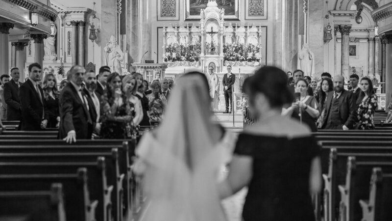 Bride being escorted down the aisle by her mother in a grand Catholic church, capturing a heartfelt and traditional wedding moment.