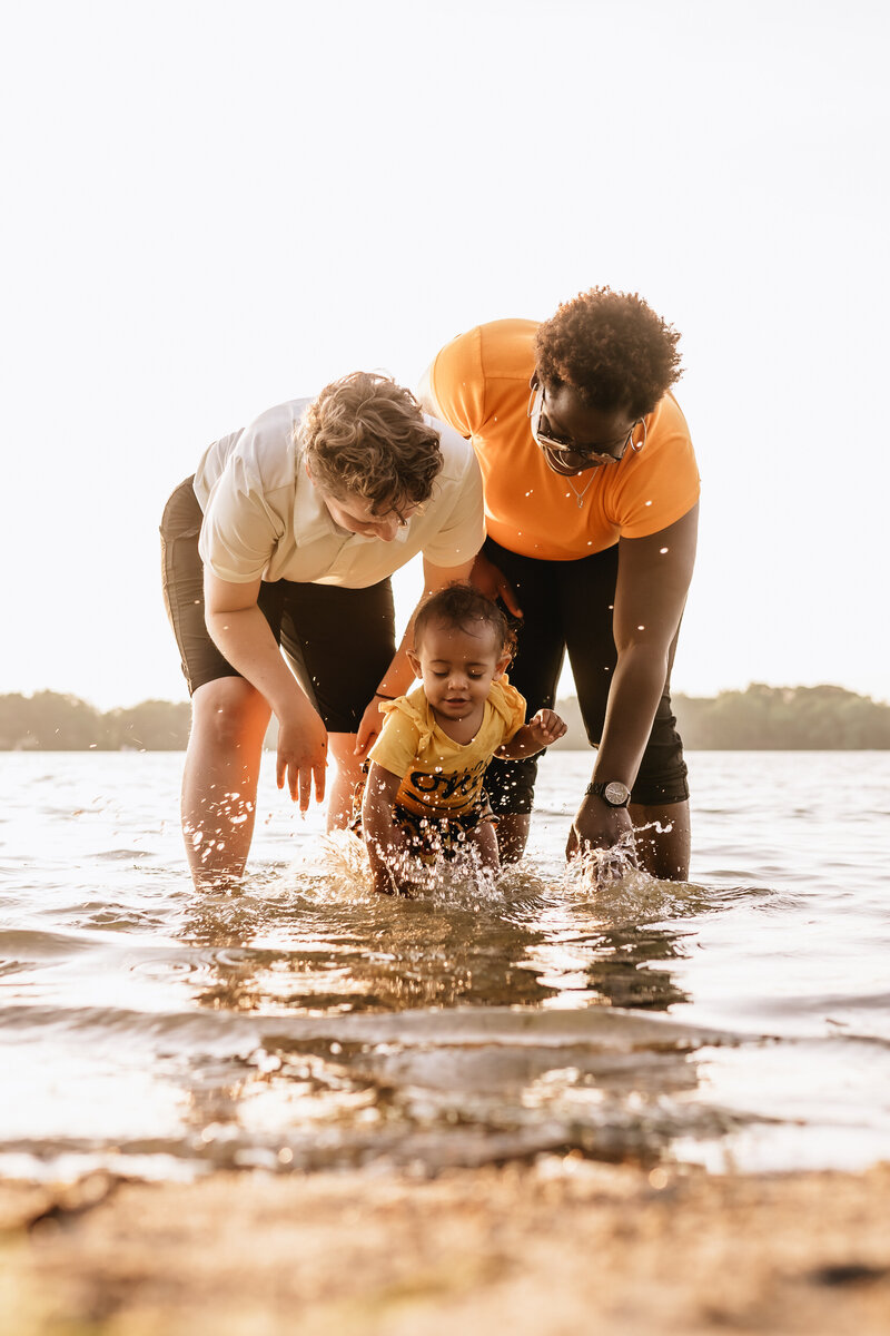 A family of three stands in shallow water at the edge of a lake.