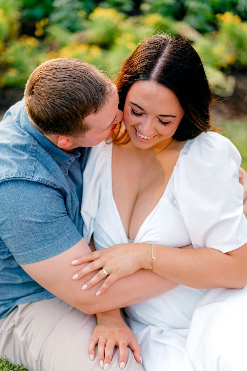 A man whispering into his future bride's ear as she smiles ear to ear.