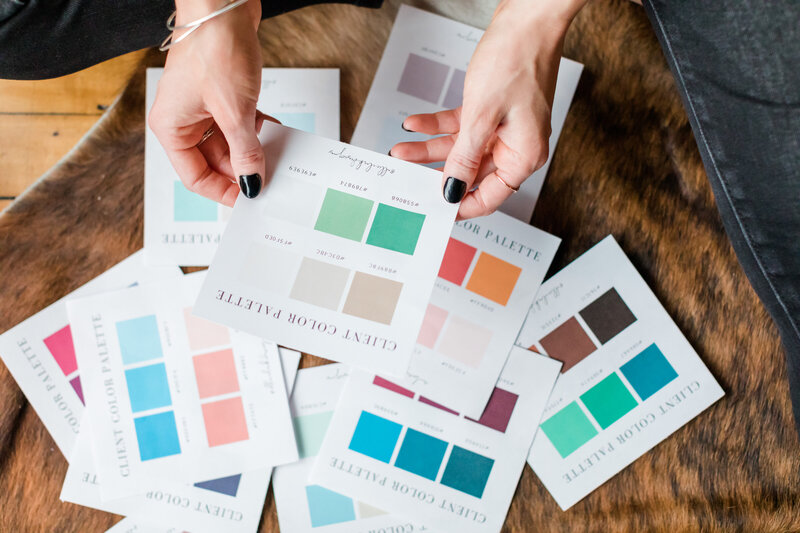 Overhead image of Allie Clark seated on the floor, carefully arranging printed client color palettes on a cowhide rug. A cozy mug of coffee and natural wood table create a warm, grounded environment — reflecting the thoughtful, hands-on nature of the Base Collection branding process.