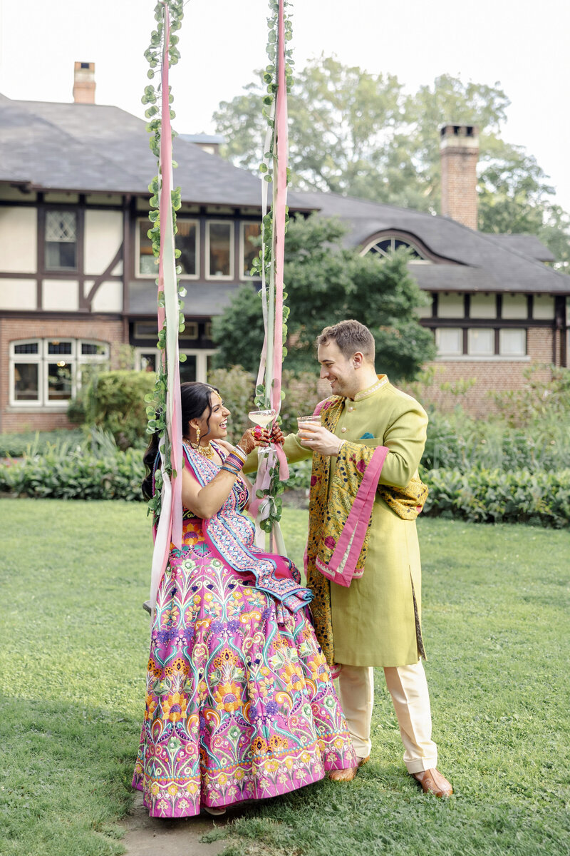 Newlyweds in colorful traditional clothing toast with cocktails in a garden. The Indian woman is on a swing decorated with ribbons and flowers. A large, charming house is in the background.