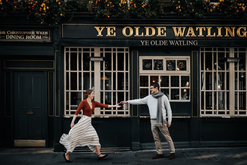 Couple-wearing-vintage-outfit-running-in-front-of-iconic-london-pub-during-pre-wedding-photo-shoot