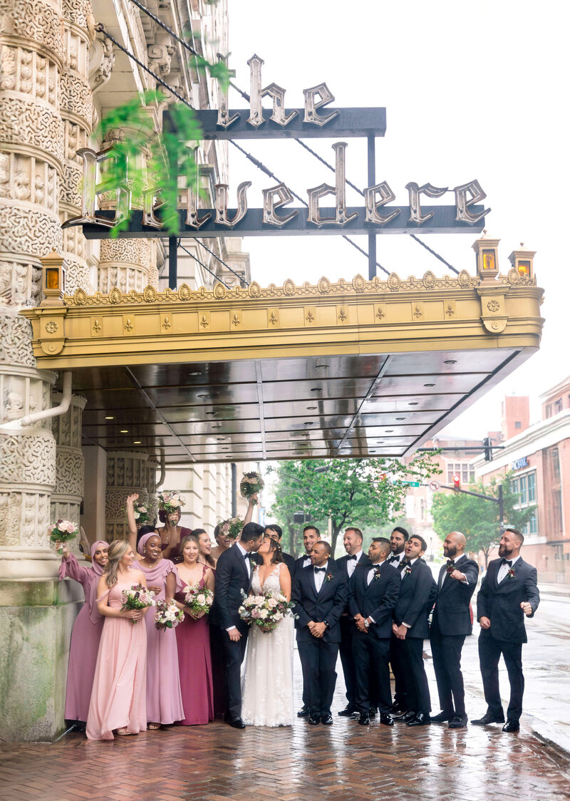 A wedding party poses outside The Belvedere. The couple stands in the center, surrounded by bridesmaids in pink and purple dresses and groomsmen in tuxedos. Everyone is smiling and celebrating under the ornate building entrance.