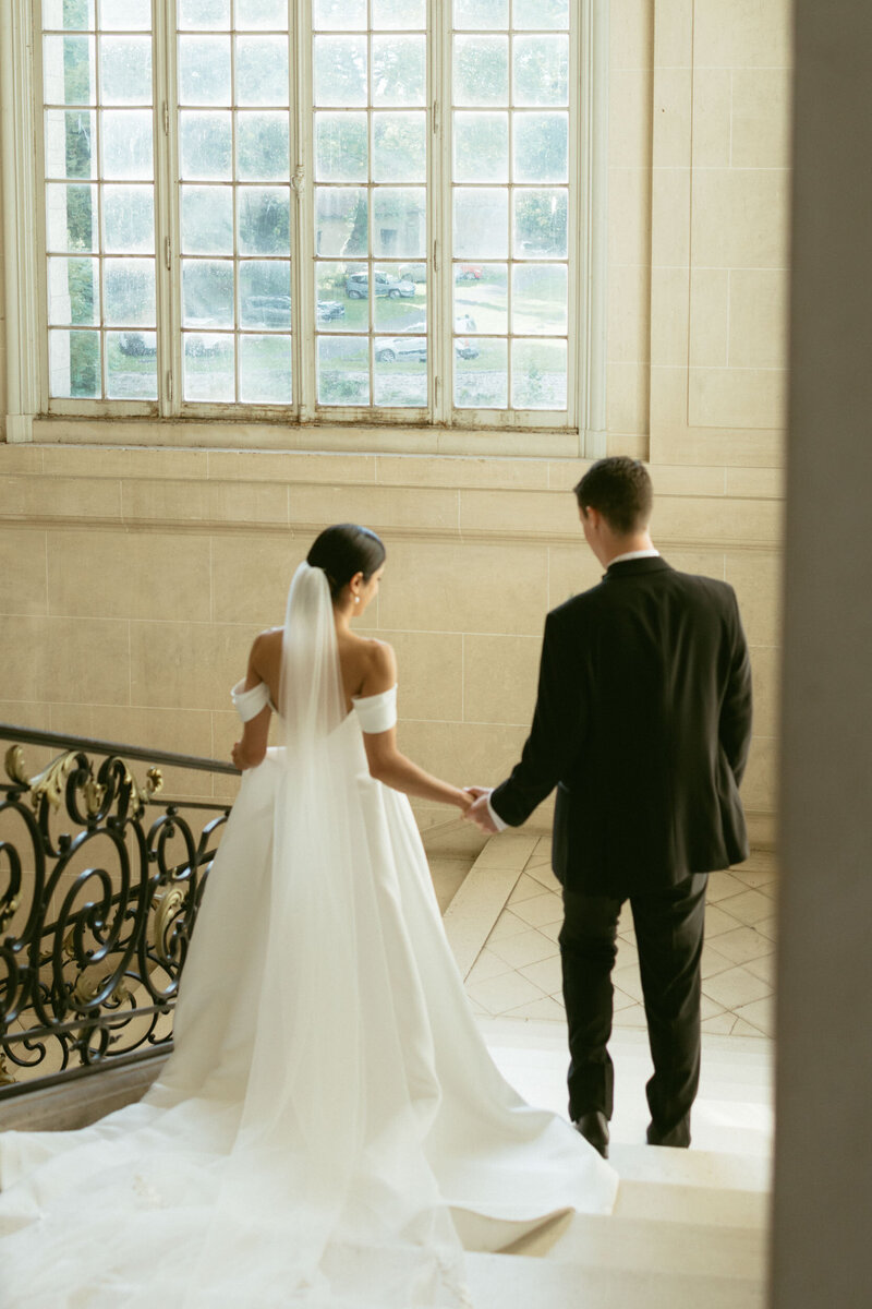 A bride and groom walk down the grand staircase of a French chateau hand in hand.