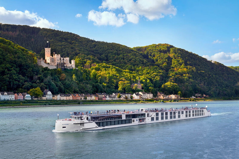 A modern white river cruise ship glides along a wide river, with a hillside dotted with trees and a historic castle perched above a small village in the background under a partly cloudy sky.