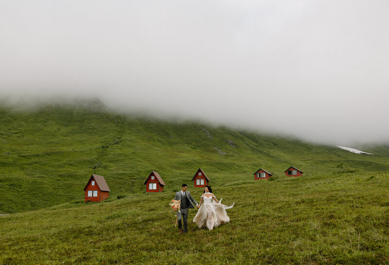 Couple holding hands and running down a hillside at Hatcher Pass, Alaska, with red A-frame cabins and fog rolling over the mountains behind them.