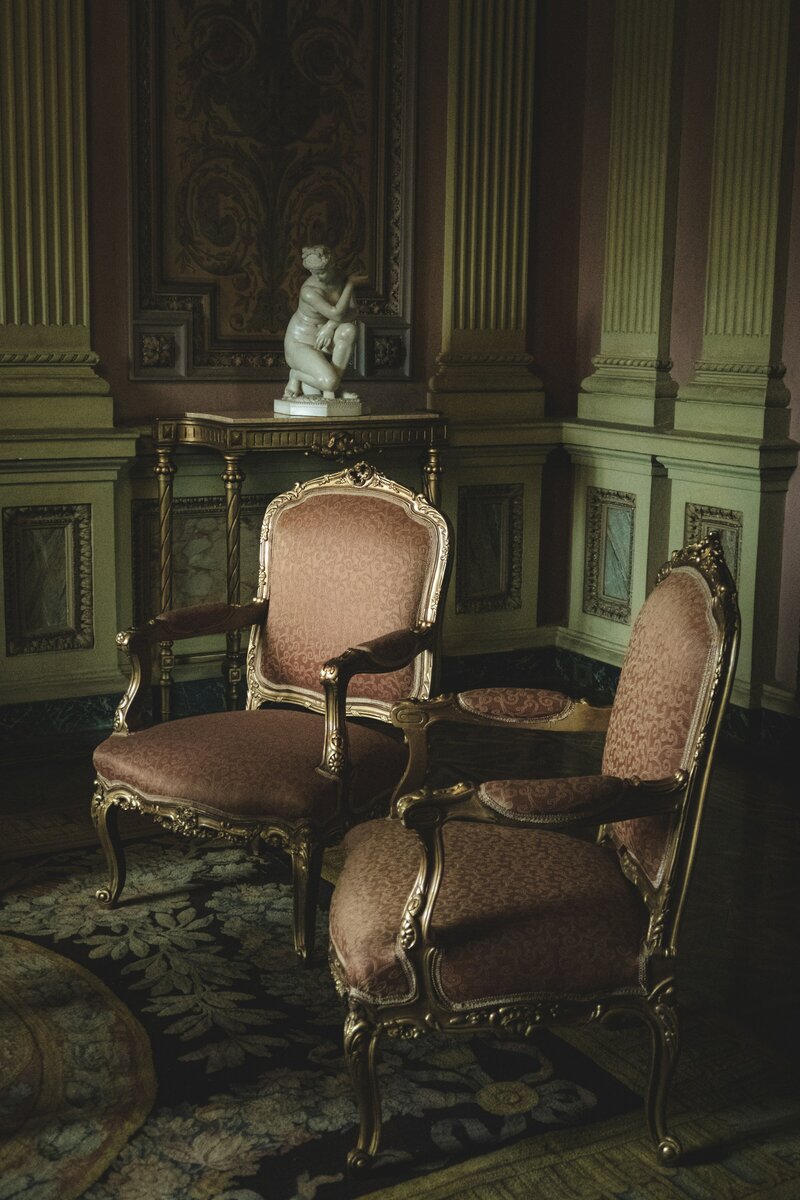 Zwei elegante antike Sessel mit goldenen Rahmen und gemustertem Stoff, platziert in einem prächtigen italienischen Palazzo mit hohen Säulen, verzierten Wänden und einer Marmorstatue auf einem dekorativen Tisch.