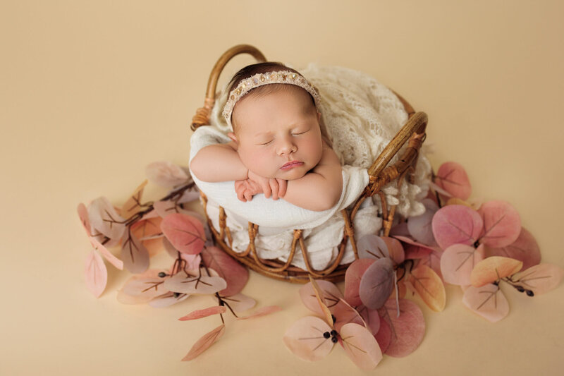 A newborn baby is peacefully sleeping, curled up on a soft, dusty rose-colored background. The baby is naked, showcasing their delicate, natural form with adorable baby rolls. A floral headband with pink and green flowers adorns the baby's head, adding a touch of sweetness to the serene scene. The baby's tiny hands are gently placed near their face, and their expression is calm and content, creating a heartwarming and tender portrait.