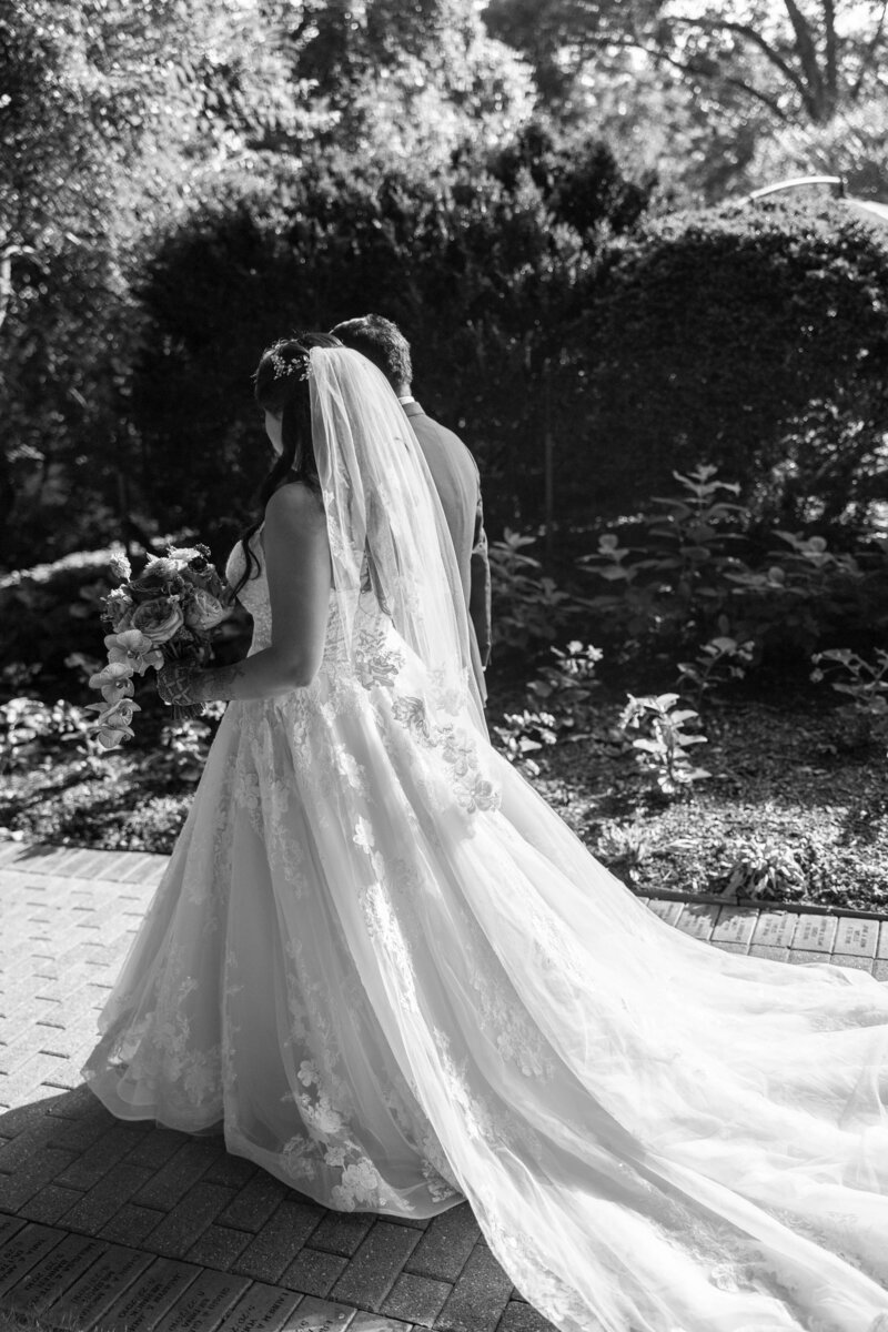 A black and white photo of a bride and groom walking away down a garden path. The bride is in a long lace gown with a veil, holding a bouquet. The groom is in a suit, and they are surrounded by lush greenery.